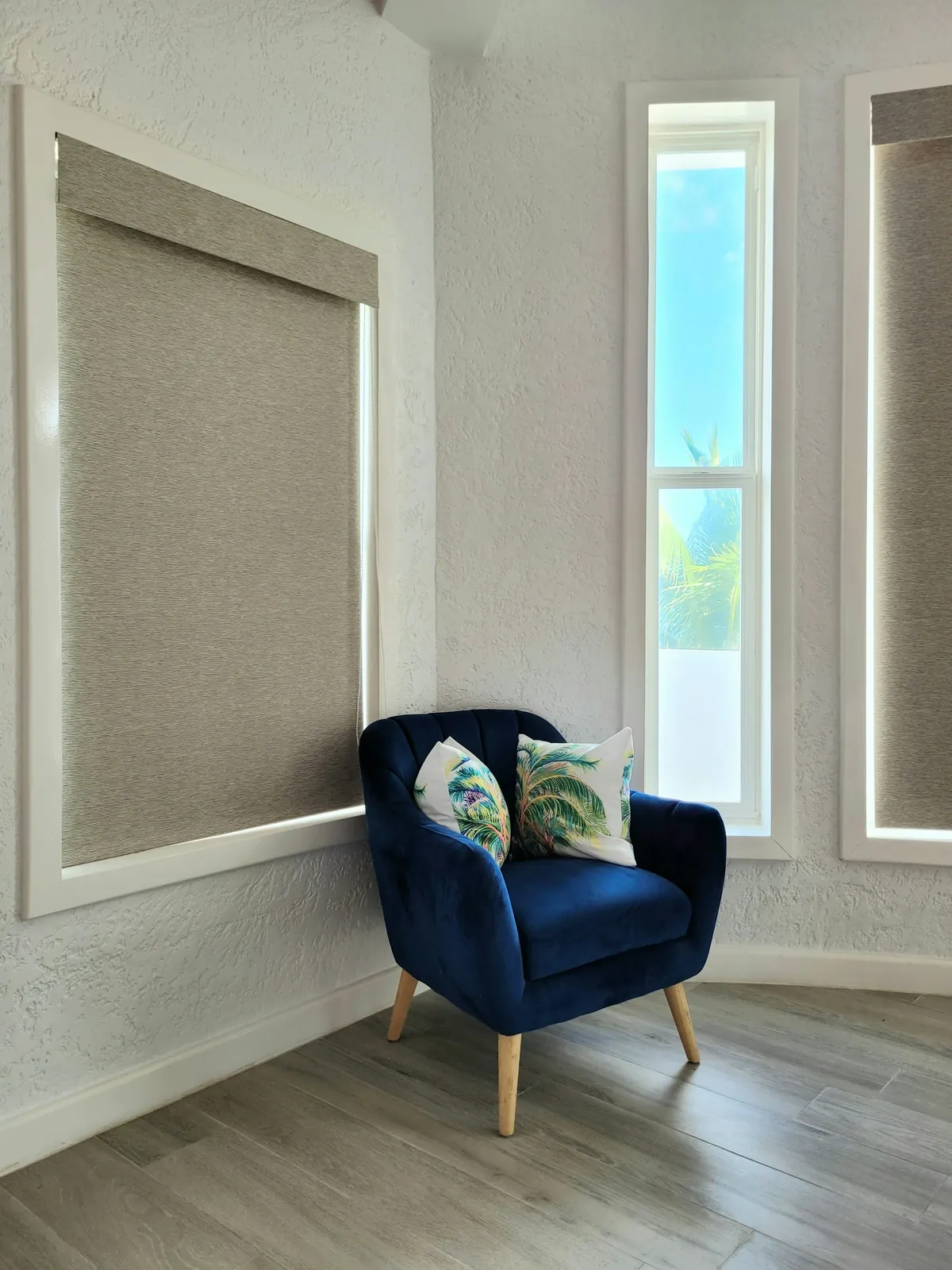 Woven roller shades over tall windows with a blue velvet armchair below