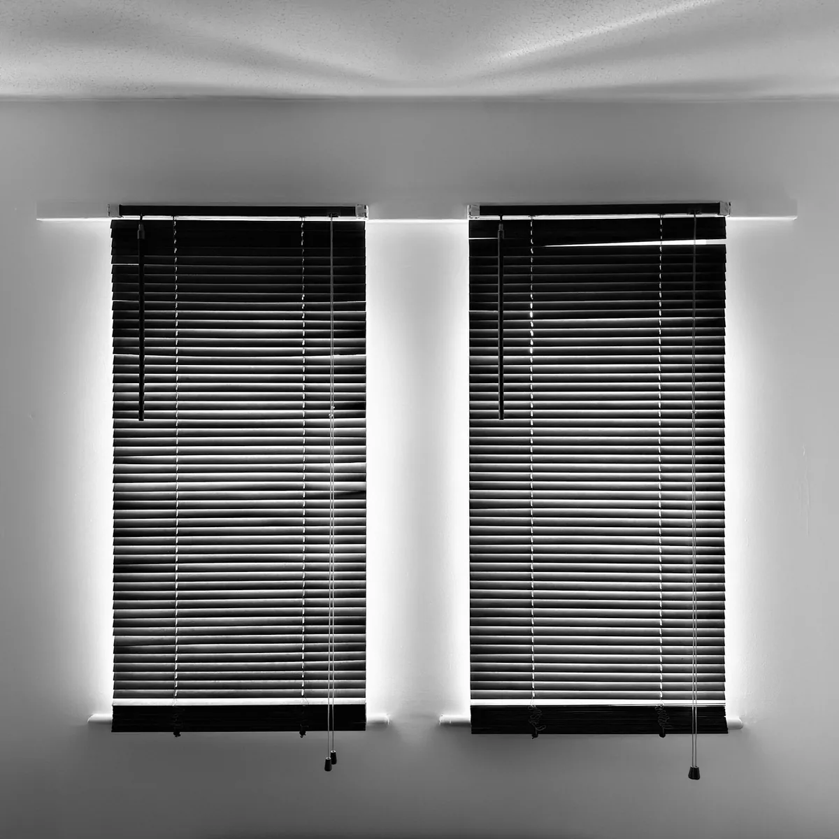 Two windows with dark horizontal blinds lowered, light glowing round the edges