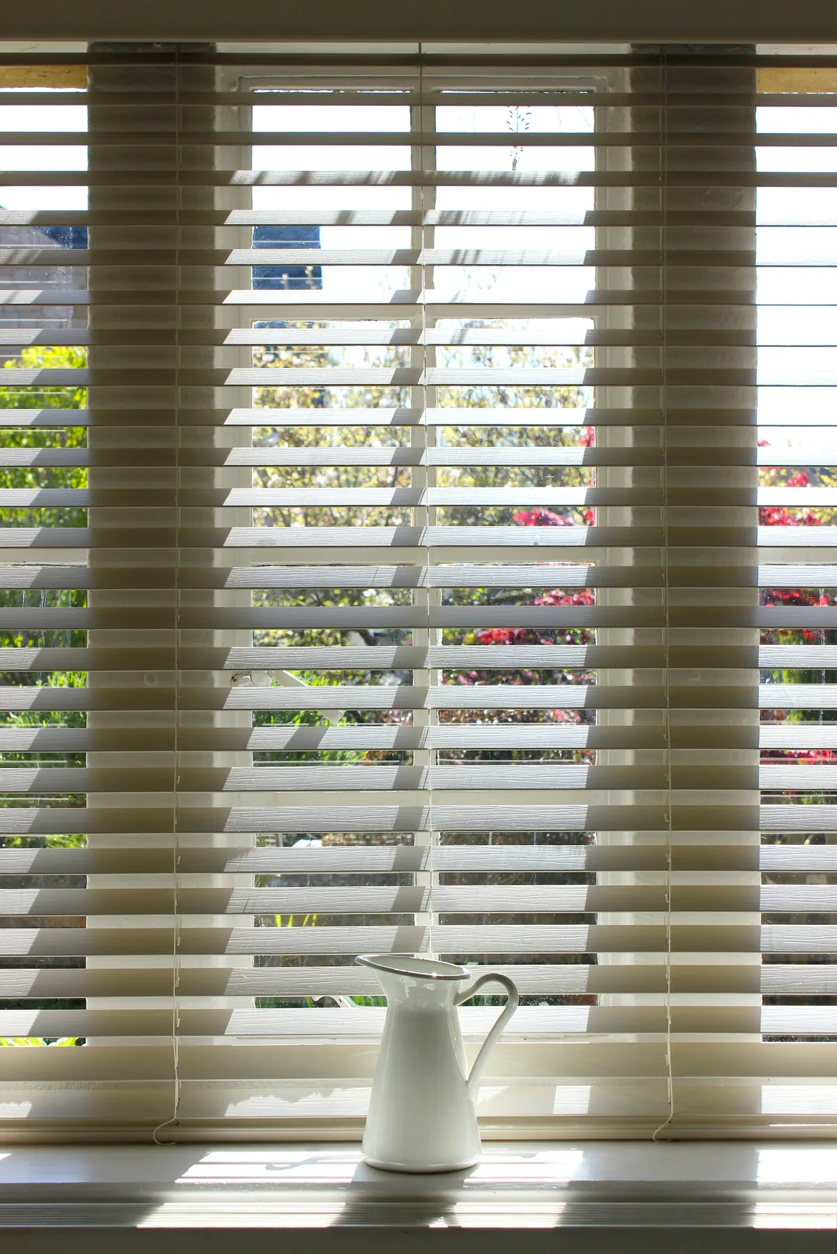 White wood blinds on tall windows with a white jug on the sill