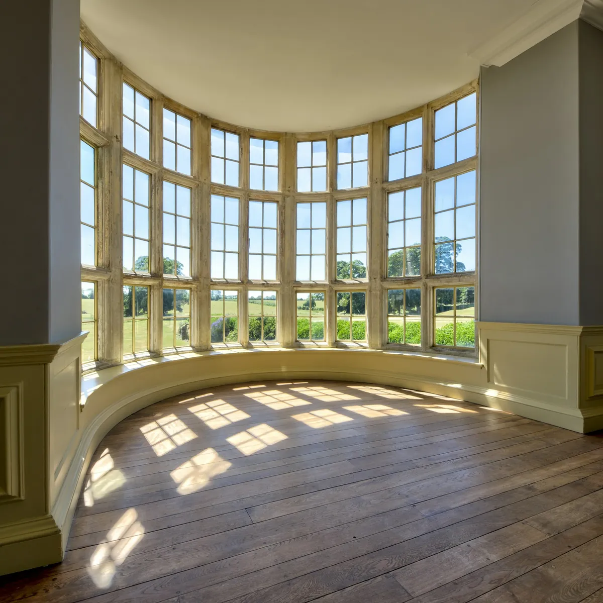 Curved bay of tall windows with sunlight across a wood floor